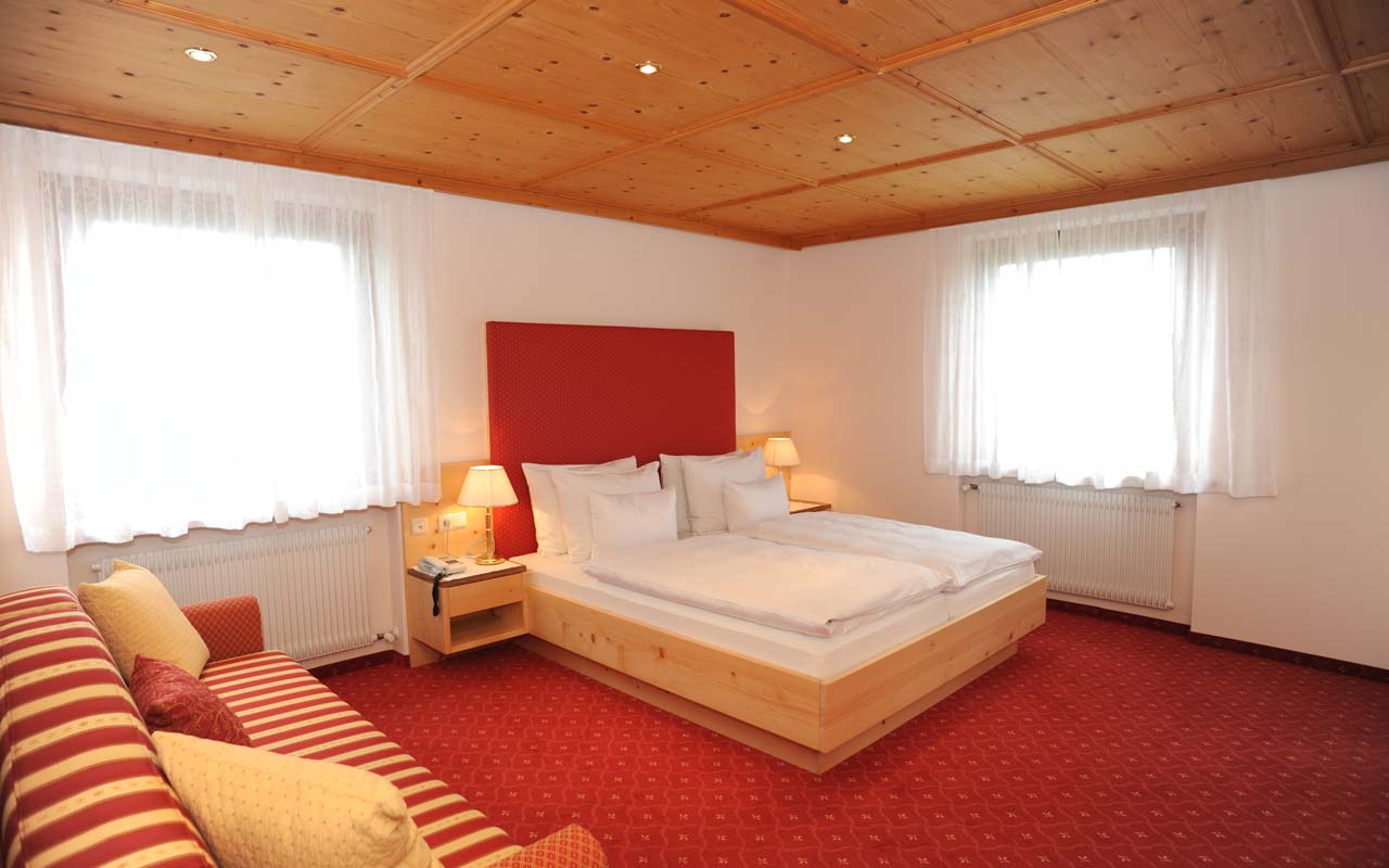 Ein großes  Zimmer mit rotem Teppichboden und zwei Fenster mit weißen Vorhängen wobei sich im linken Bereich ein großes  rot-weiß gestreiftes Sofa befindet und auf der rechten Seite ein Doppelbett mit weißer Bettwäsche und zwei Nachtkästchen mit zwei Nachttischlampen stehen
