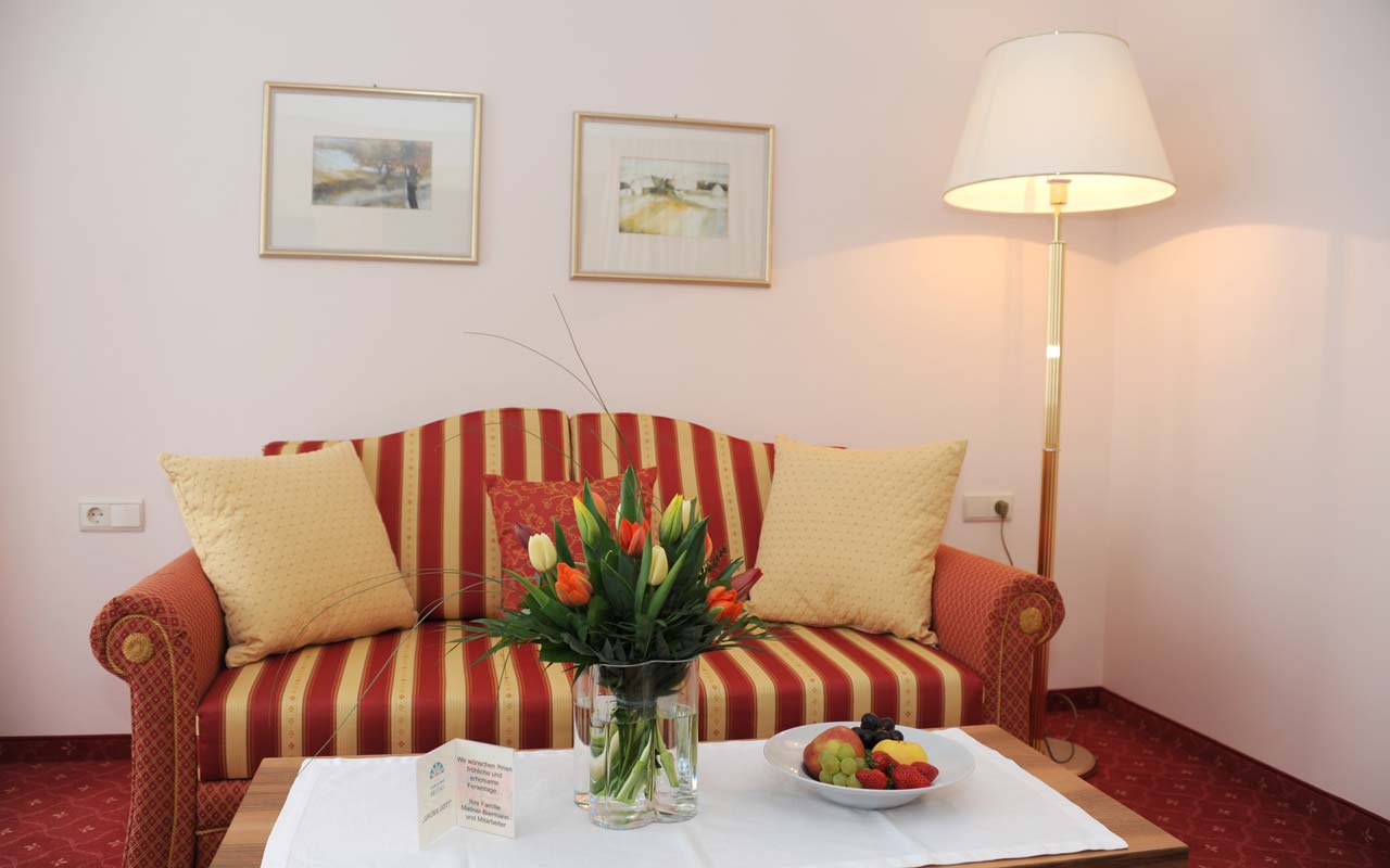 Raum in dem ein großes rot-gelb gestreiftes Sofa steht mit einem Sofatisch mit weißem Tischtuch, ein Blumenstrauß und eine Obstschale; und rechts daneben steht eine weiße Lampe wobei an der Wand, hinter dem Sofa zwei Bilder hängen