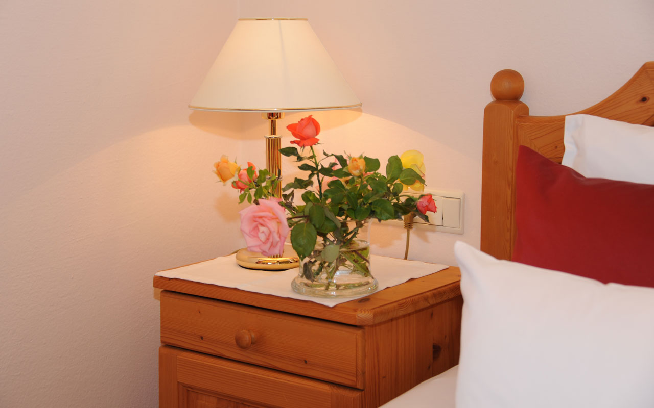 Ein kleiner Nachttisch auf dem sich ein Blumenstrauß mit rotem, gelben, rosanen und Orangen Blumen steht und eine weiße Nachttischlampe und rechts daneben sieht man ein Teil eines Bettes mit roten und weißen Kissen
