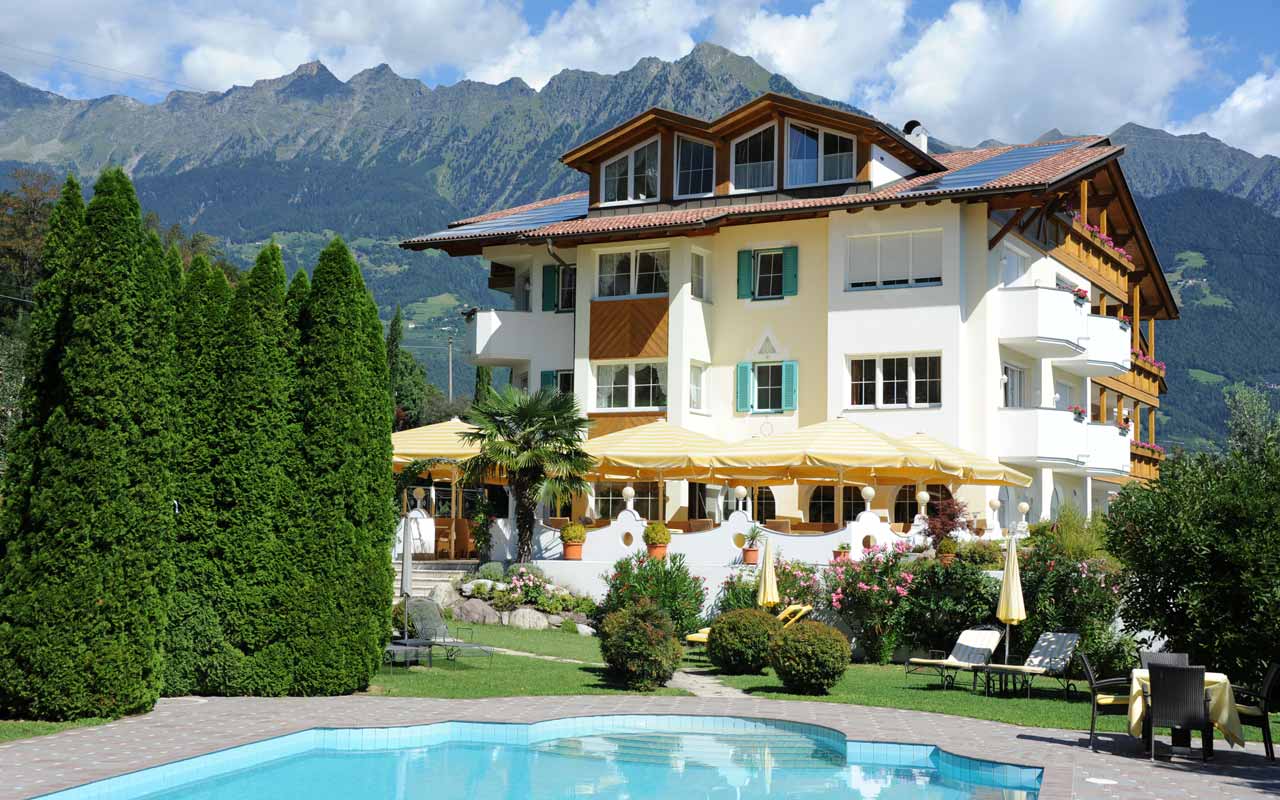 Es ist das Hotel Kristall mit Terrasse zu erkennen wobei davor eine Wiese mit Schwimmbad und Bäume stehen und dahinter befinden sich Berge