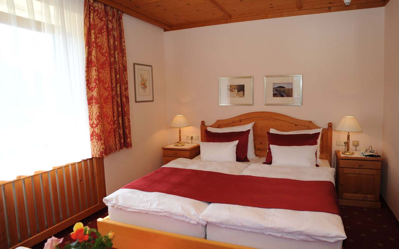 Hier ist ein helles Zimmer mit einem großem Fenster und roten Vorhängen, ein Doppelbett mit weißer und roter Bettwäsche, braune Holznachtkästchen auf denen weiße Nachttischlampen platziert sind und an den Wänden drei Bilder zu sehen