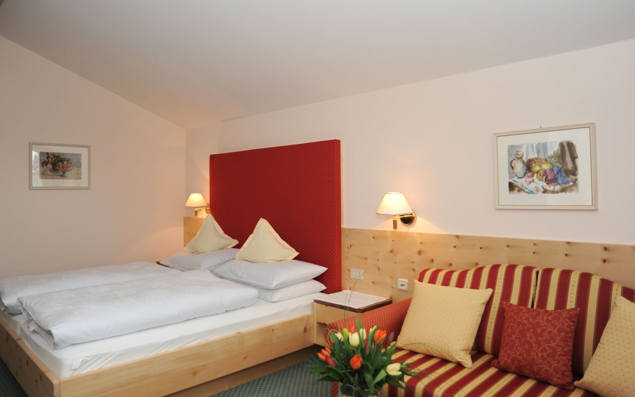 Auf der linken Seite befindet sich ein Doppelbett mit weißer Bettwäsche und an der Wand hängt ein Bild und auf der rechten Seite befindet sich ein rot-gelbgestreiftes Sofa und ein Sofatisch auf dem ein Blumenstrauß steht wobei an der Wand oberhalb des Sofas ein Bild hängt
