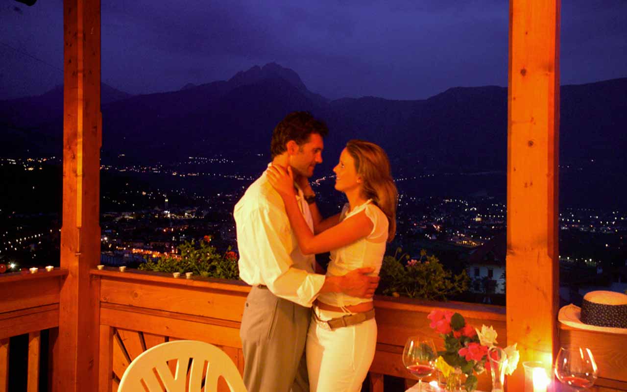 Am späten Abend steht ein Liebespaar an einem Balkon wobei sich das Paar umarmendmend in die Augen schaut und im Hintergrund sieht man die Aussicht übers Tal wobei die Lichter der Häuser in der Dunkelheit läuchten