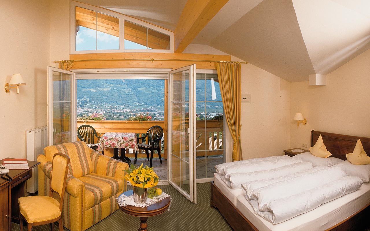 Ein Zimmer mit großem Balkon und einem grünen Teppichfuß boden wobei auf der linken Seite ein großes  Sofa, ein Sofatisch mit gelben Blumen, ein Stuhl und ein Schreibtisch stehen und auf der rechten Seite ein Doppelbett mit weißer Bettwäsche stehen
