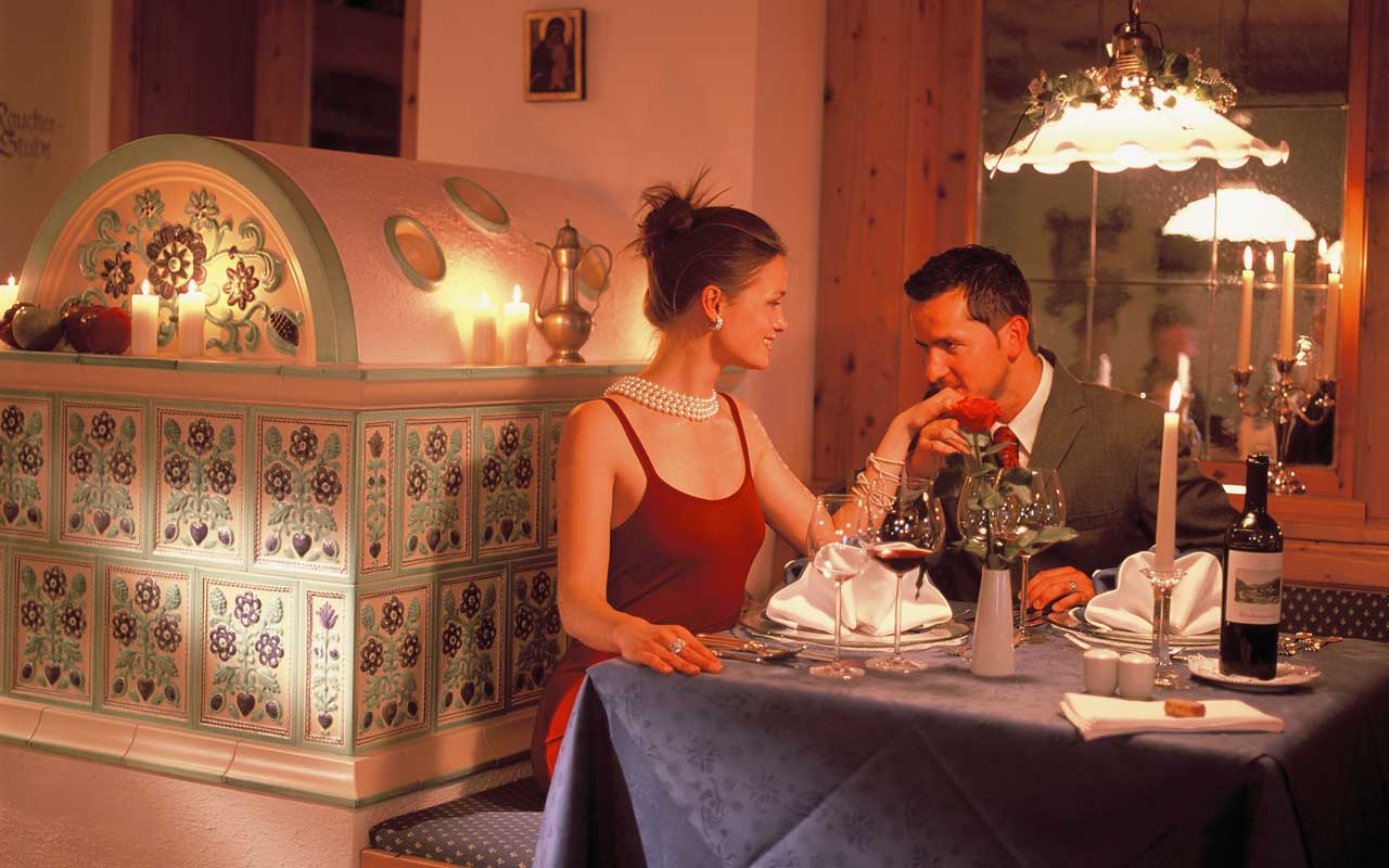 In einer Stube neben einem Kachelofen, sitzen eine Frau mit rotem Kleid und einer Perlenkette um den Hals und ein Mann mit grauen Anzug auf einer Eckbank am Esstisch, wobei der Mann der Frau einen Handkuss übergibt