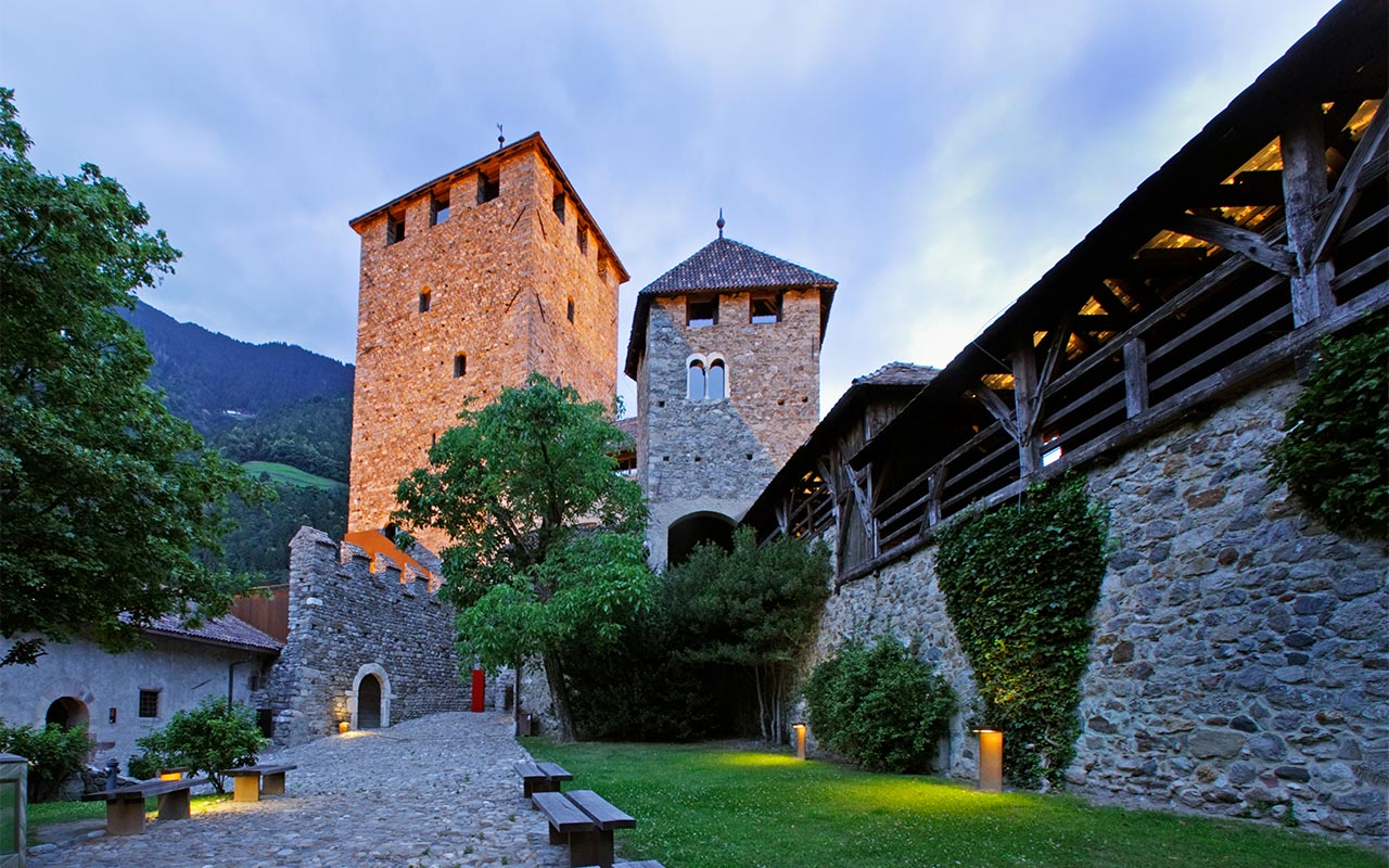 Im Abendlicht ist im Hof von Schloss Tirol das Schloss zu sehen wobei sich rundherum grüne Bäume und Wiesen befinden