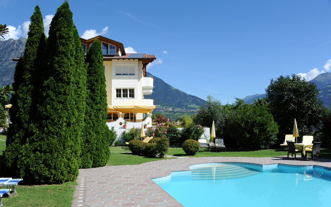 Unter strahlend blauen Himmel ist das Hotel Kristall zu erkennen mit dem Garten davor worauf sich viele Bäume, das Schwimmbad und Sonnenliegen befinden