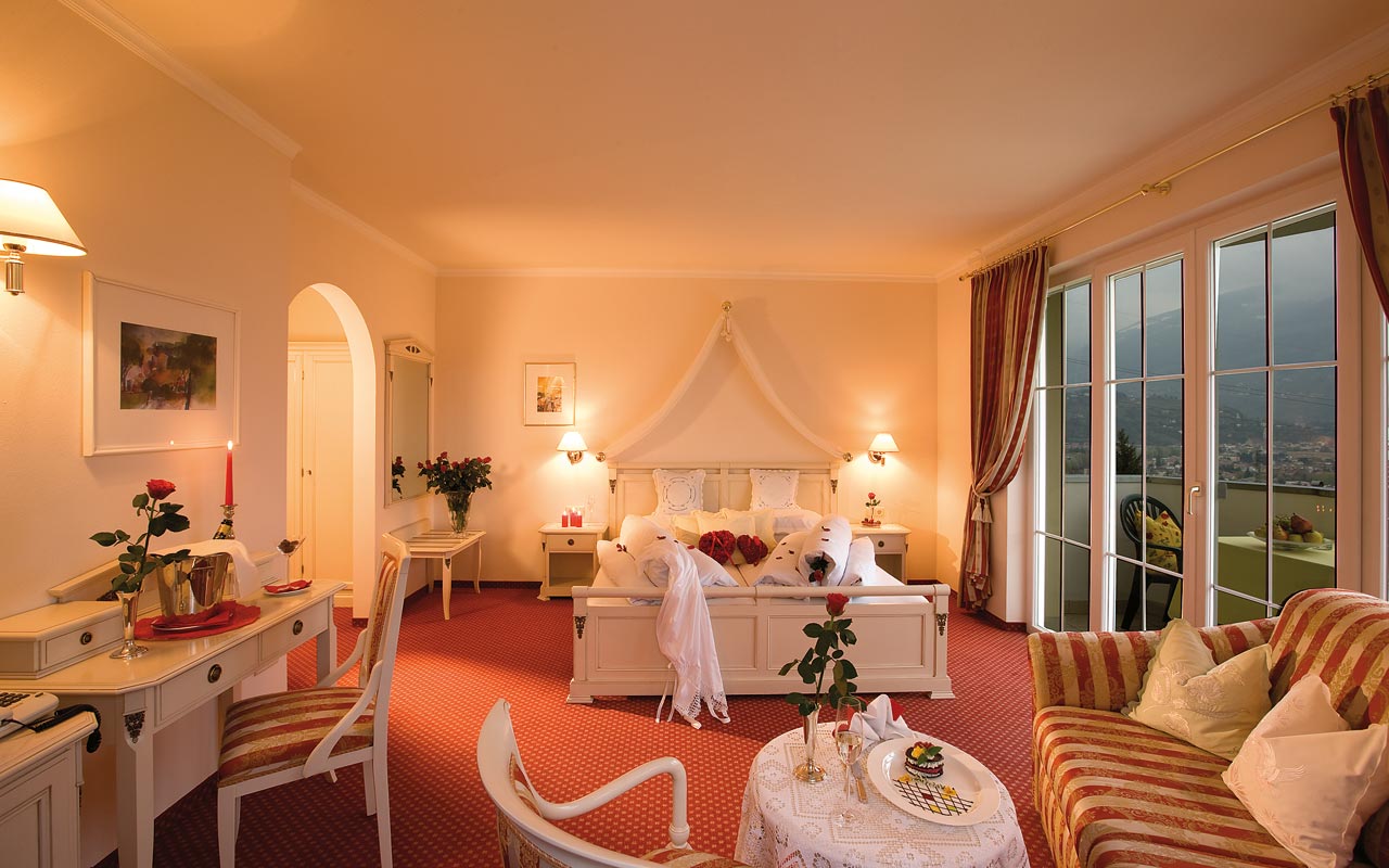 Ein großes  helles Zimmer mit einem großen weißem Bett, einem gold-orangenen Sofa auf der rechten Seite, einem Tischchen mit einem Stuhl auf der linken Seite und einem Balkon mit Talblick
