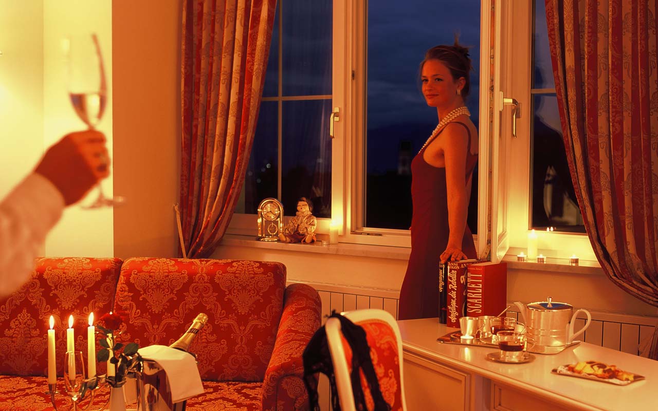 In einem Zimmer steht eine Frau mit einem rotem Kleid mit dem Rücken zum offenen Fenster geneigt, wobei sich links neben ihr eine rote Couch mit einem kleinen Tisch auf dem eine Sektflasche und leuchtende Kerzen stehen und rechts von ihr ein Stuhl und ein Schreibtisch auf dem Bücher und Tee sind befinden