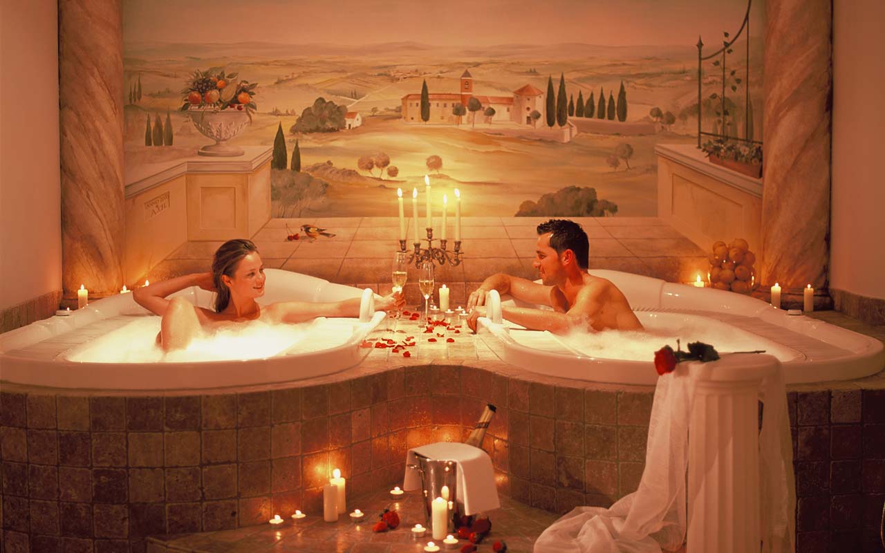Eine Frau und ein Mann die in einem Wellnessbereich sind und jeder befindet sich in einem Whirlpool, gefüllt mit Badeschaum und rundherum sind Kerzenlichter, Rosenblätter und eine gekühlte Sektflasche